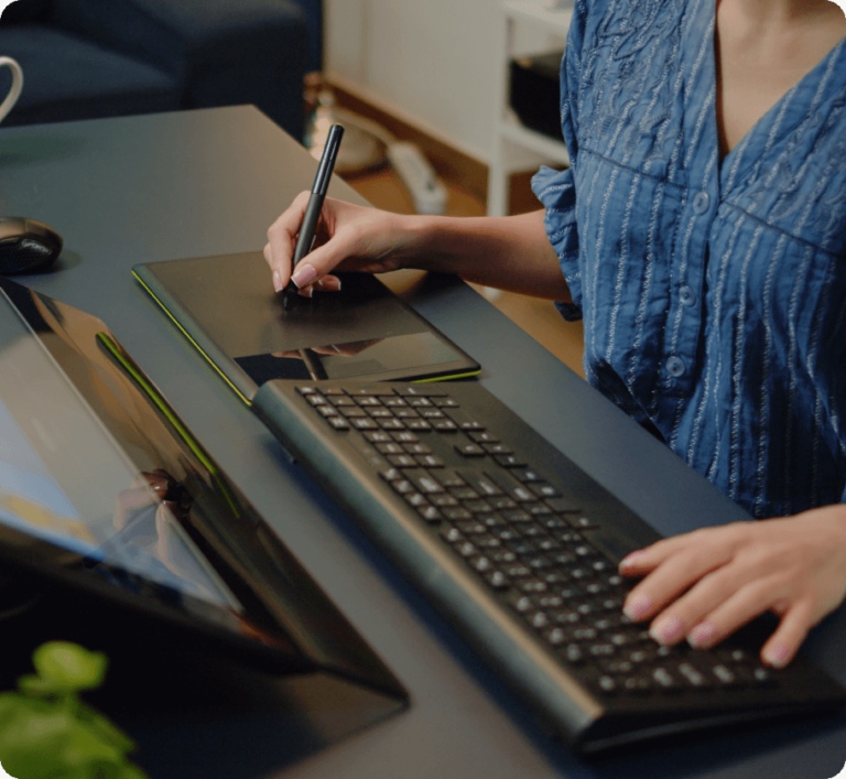 A digital artist working on a drawing tablet, using a stylus pen while operating a keyboard in a modern workspace.