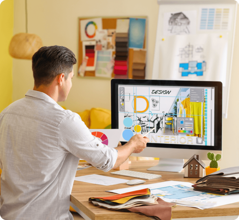 A designer working on an interior design project at a desk, using a computer with design software open, surrounded by color palettes and sketches.